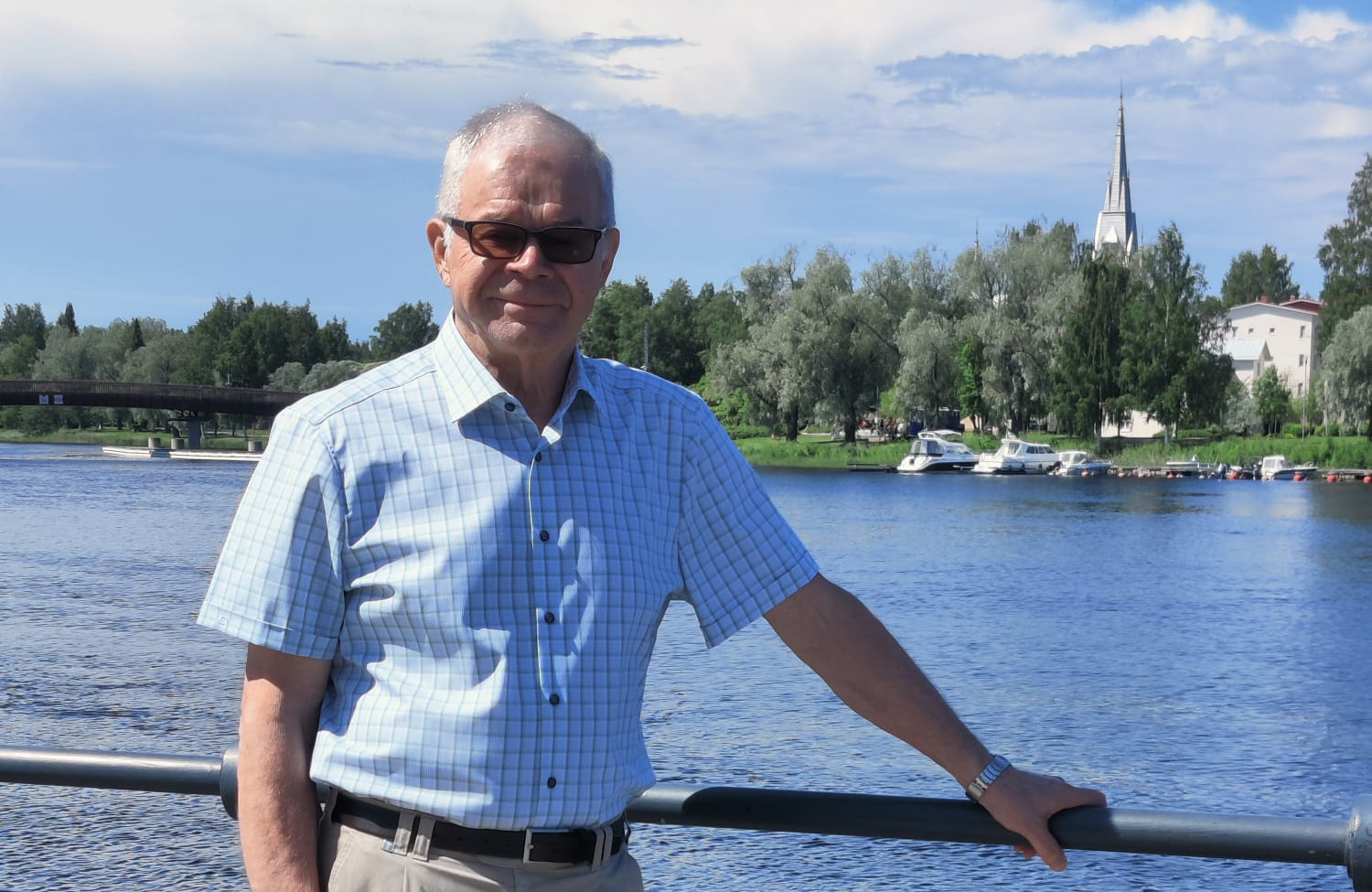 Aurinkolasipäinen mies nojaa toisella kädellään kaiteeseen, taustalla jokea, jonka vastarannalla veneitä ja...