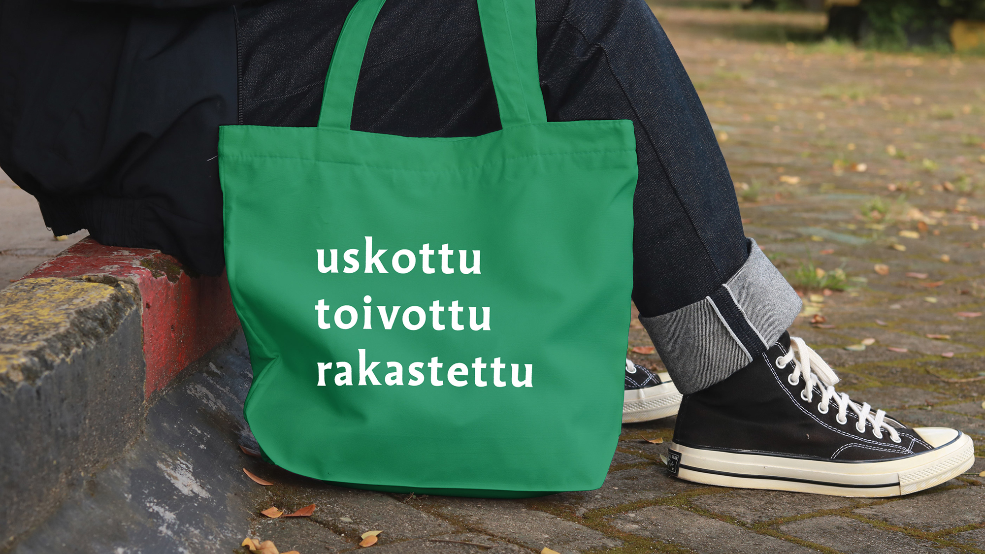 Vihreä kassi tekstillä uskottu, toivottu, rakastettu, taustalla istuu ihminen.