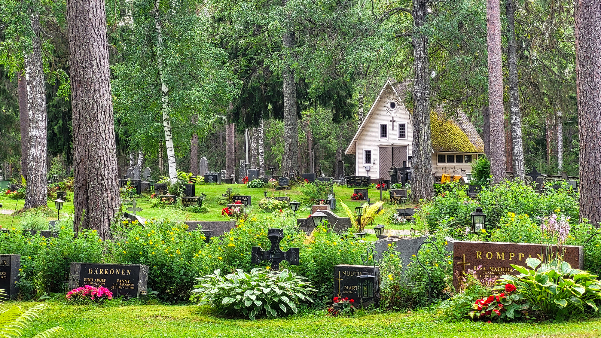 Enon hautausmaalla on hautakiviä ja kukkaistutuksia. Niiden lomassa kasvaa puita. Taustalla näkyy rakennus.