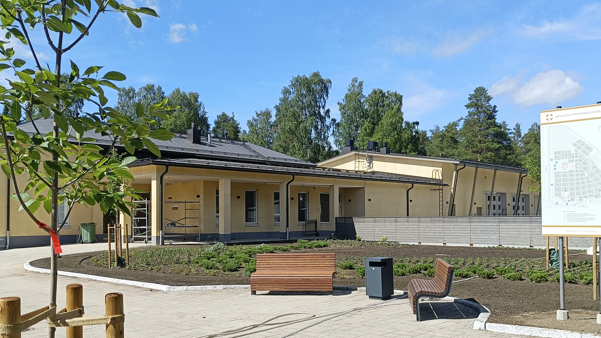 Taka-alalla on hautausmaan uusi keltainen toimisto- ja huoltorakennus. Etualalla on istutuksia ja penkkejä.
