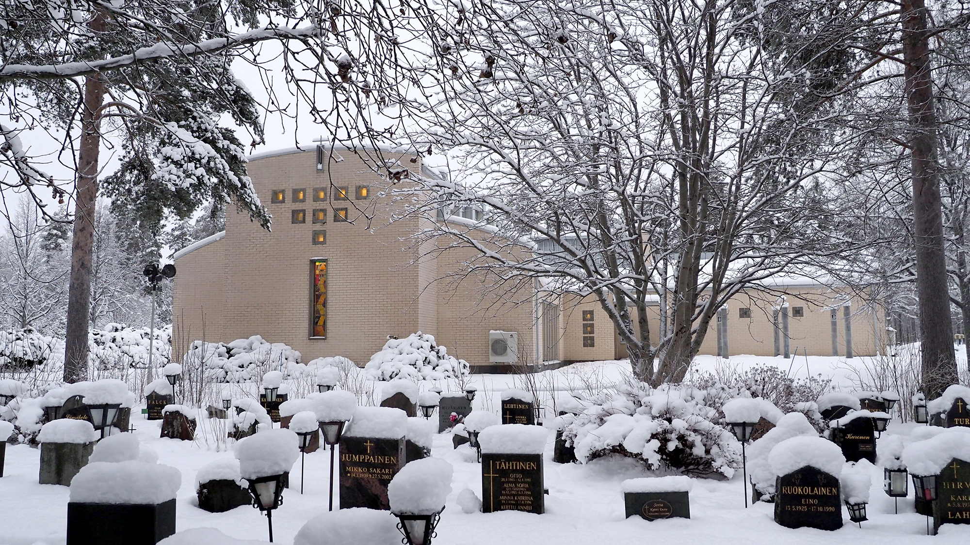 Ristinkappeli kuvassa taustalla, etualalla hautausmaata ja hautakiviä, joiden päällä on lunta.