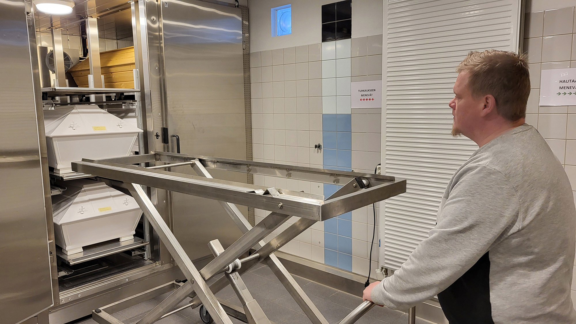 Pasi Tarnanen hauta-arkkujen kylmäsäilytystilojen ääressä kuljetustelineen kanssa.