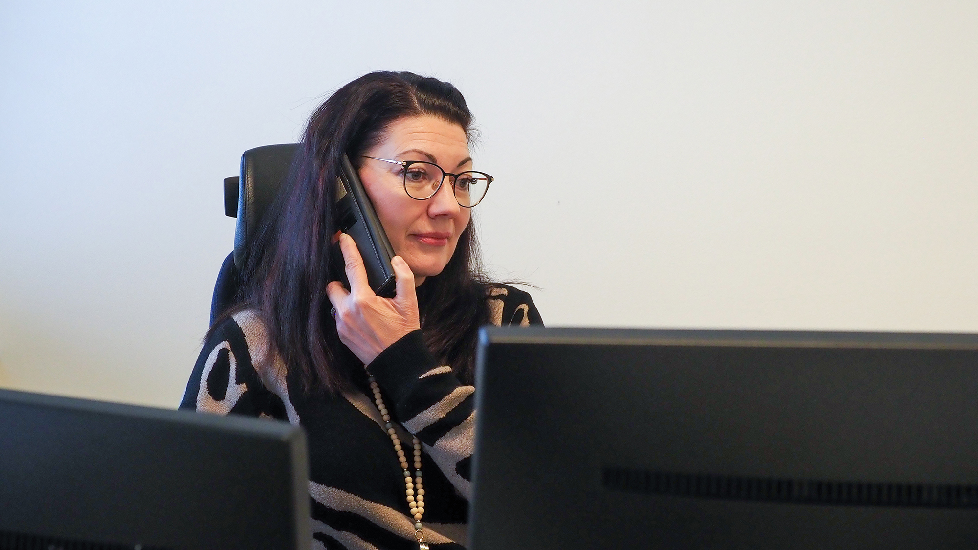 Päivi Hirvosella on puhelin korvallaan. Hänen edessään on kaksi tietokoneen näyttöä.