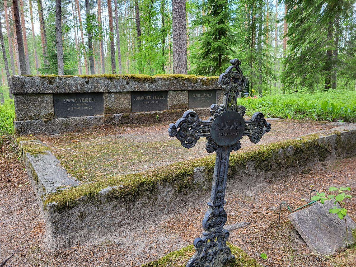 Hautakiviä, puita, edustalla rautainen koristeellinen risti.