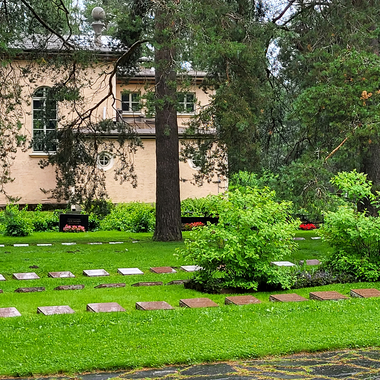 Muistokiviä nurmikolla hauta-alueella, taustalla puita ja vaalea rakennus.