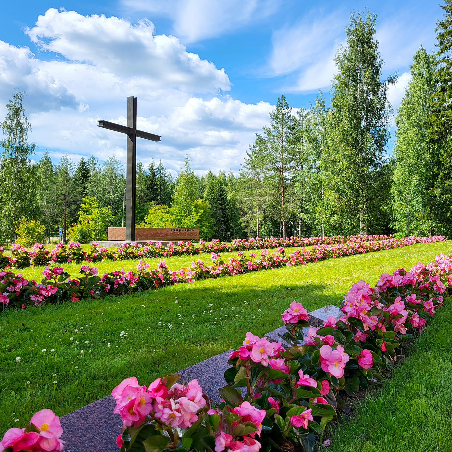 Kukkaistutuksia ja hautakiviä etualalla, taustalla suuri risti ja muistomerkki.