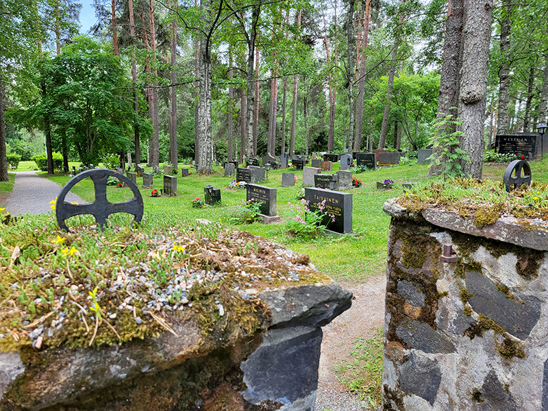 Kivinen aita ja portti, jonka päällä rautainen ristikoriste, taustalla puita, nurmea ja hautakiviä.