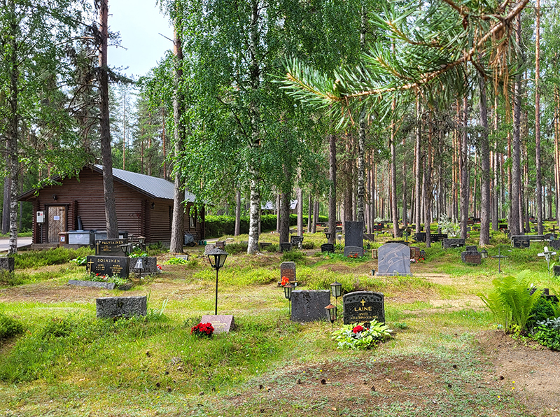 Metsäinen hautausmaa, ruskea huoltorakennus vasemmalla.