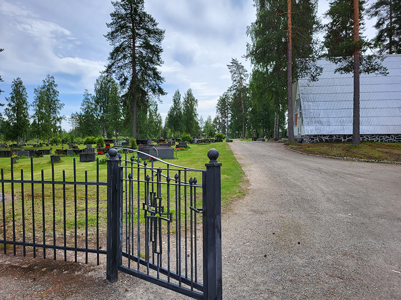 Tuupovaaran hautausmaa Rautainen aita ja portti, taustalla vaalea rakennus ja hautausmaa-alue.