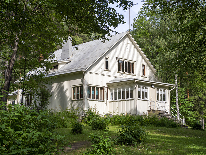 Männikköniemen kesäkoti Kesäinen kuva, ruohoa, puita ja pensaita, auringon valoa pienessä rinteessä. Rinteen päällä vanha, puinen ja vaalea kaksikerroksinen rakennus, jossa etuosassa veranta, jonne noustaan kivirappusia myöten.