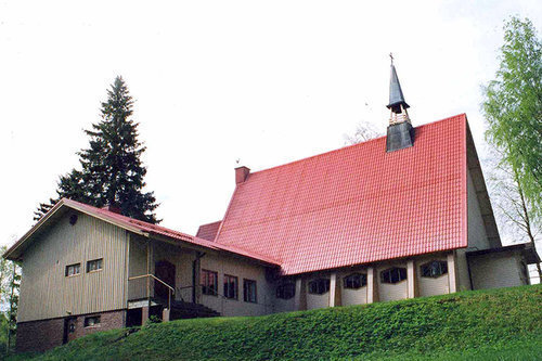 Kesäinen kuva, jossa etualalla nurmikkoinen rinne ja sen päällä vaalea rakennus, jossa toisessa osassa korkea punainen katto sekä pieni kellotorni. Oikealla matala lisäsiipi. Puita taustalla.