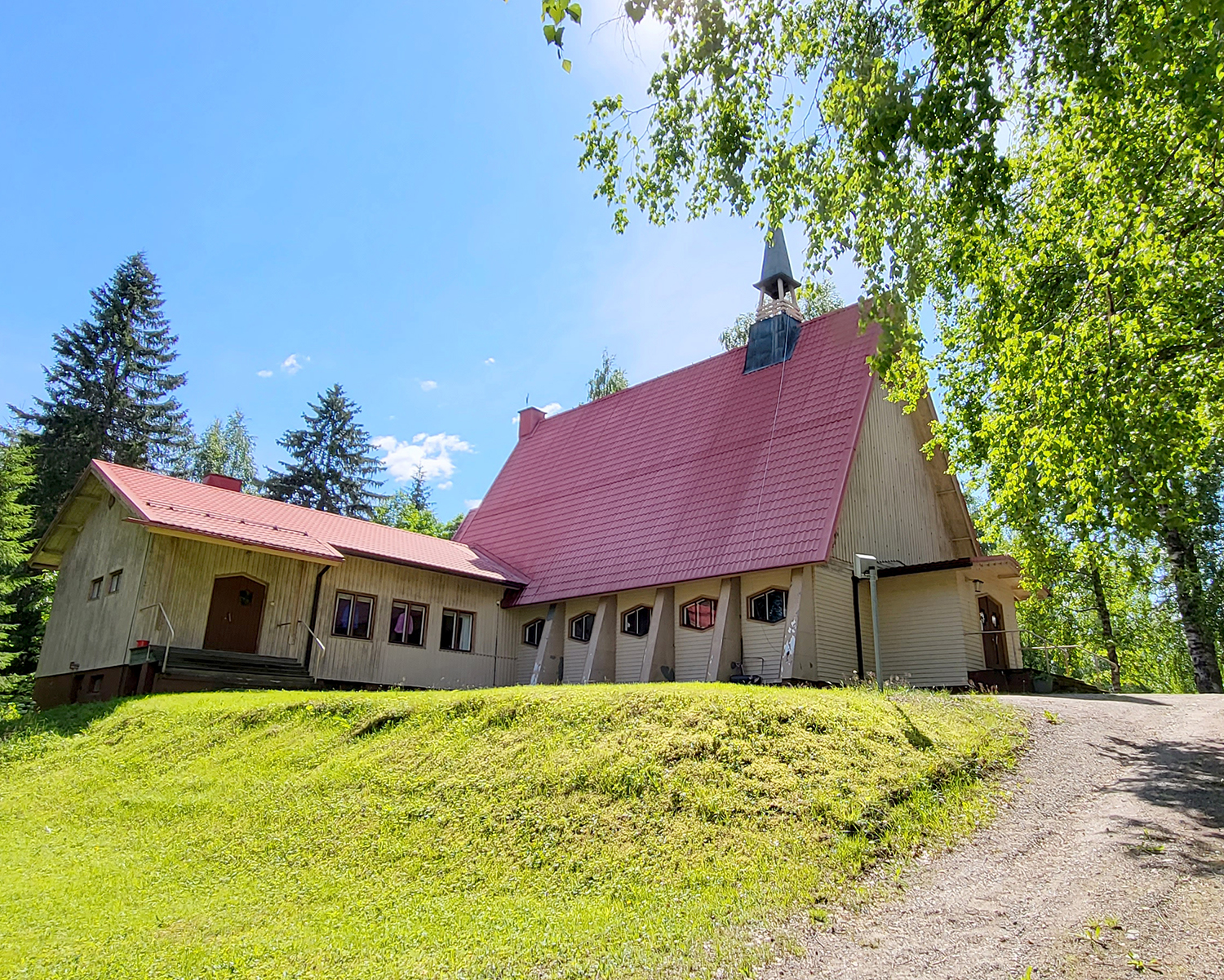 Kesäinen kuva, jossa etualalla nurmikkoinen rinne ja sen päällä vaalea rakennus, jossa toisessa osassa korkea punainen katto sekä pieni kellotorni. Oikealla matala lisäsiipi. Puita taustalla.