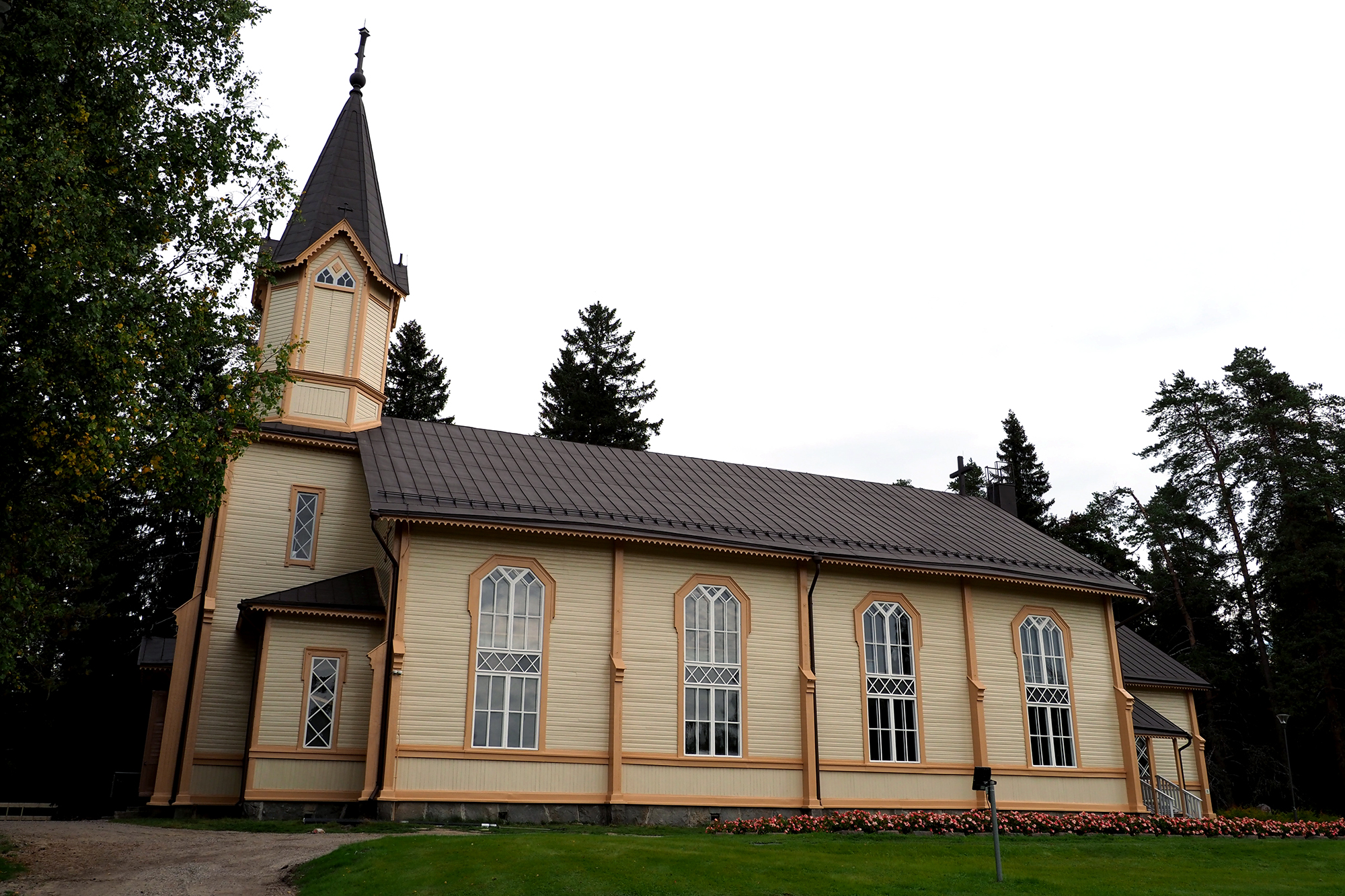 Kesäinen kuva, jossa kirkkorakennus kuvattu sivusta. Rakennus on vaalea ja siinä on neljä suurta sivuikkunaa. Rakennuksen etuosassa on kolmikerroksinen, suippokattoinen torni, jossa katon päällä risti. Havupuita rakennuksen taustalla.