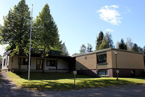 Kesäinen kuva matalasta keltatiilinsesta talosta, jonka edessä parkkipaikka, lipputanko ja kaksi puuta.