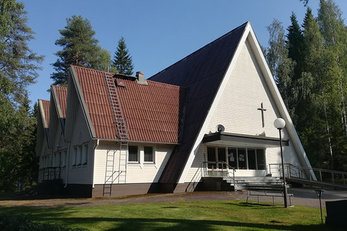 Kolmikulmainen rakennuksen talonpääty, jossa risti sisäänkäynnin yläpuolella. Puita ja nurmikkoa ympärillä.