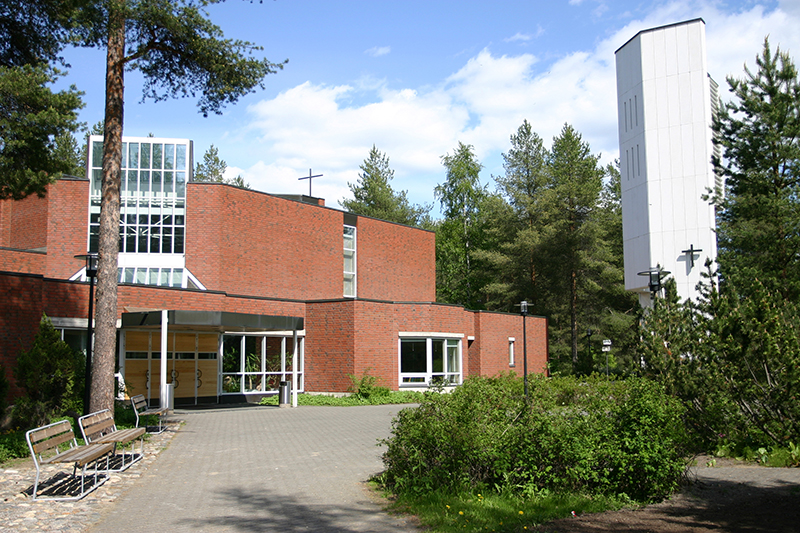 Kesäinen kuva, jossa pensaita, puistonpenkkejä, mäntyjä sekä harmaalla laatoituksella oleva piha-alue etualalla. Oikeassa laidassa korkea betoninen, valkoiseksi maalattu kellotorni, jonka seinässä pieni risti. Kuvan taustalla punatiilinen rakennus ja sen sisäänkäynti sekä ikkunoita. Yksikerroksinen rakennus kasvaa takaosastaan korkeammaksi ja sen keskellä on suuri ikkunaseinä, katolla risti.