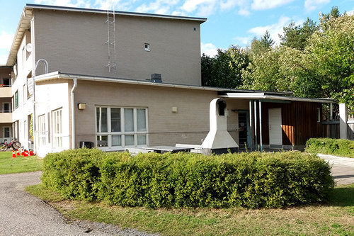 Talastupa Kesäinen kuva, jossa matalapensasaita edessä, sen takana pieni laatoitettu piha-alue, jossa korkeapiippuinen tulisija ja vaaleatiilisen kerrostalon pääty. Päädyssä yksikerroksinen erillistila ja sen sisäänkäynti.