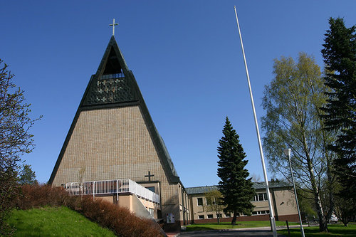 Keväinen kuva, jossa etualalla nurmikkoa ja puistoalueen pensaistoa ja puita, lipputanko ja valopylväs. Kolmikulmainen rakennuksen pääty, jonka edessä aidoitettu tasanne. Rakennuksen pääty päällystetty vaaleanruskeilla tiilillä, tumma katto luo reunuksia ja kolmion päädyn yläosaan. Päädyn yläosa avoin tila, jossa kirkonkello, ylimpänä risti. Taustalla näkyy rakennuksen kaksikerroksinen sivusiipi.