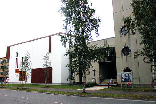 Joensuun seurakuntakeskus Kesäinen kuva, jossa autotie etualalla, pieni puistoalue nurmineen ja koivupuineen sekä jalankulkuväylä. Näiden takana rakennus, jonka oikeanpuoleinen osa harmaata betonielementtiä. Seinässä kolme pyöreää ikkunaa ja risti sekä sisäänkäyntikatos. Vasemmanpuoleinen laatikkomainen rakennus, joka pinnoitettu valkoiseksi, ruskeita puuosia ja rimoitusta alaosan ikkunoiden edessä.