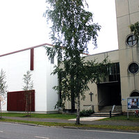 Kesäinen kuva, jossa autotie etualalla, pieni puistoalue nurmineen ja koivupuineen sekä jalankulkuväylä. Näiden takana rakennus, jonka oikeanpuoleinen osa harmaata betonielementtiä. Seinässä kolme pyöreää ikkunaa ja risti sekä sisäänkäyntikatos. Vasemmanpuoleinen laatikkomainen rakennus, joka pinnoitettu valkoiseksi, ruskeita puuosia ja rimoitusta alaosan ikkunoiden edessä.