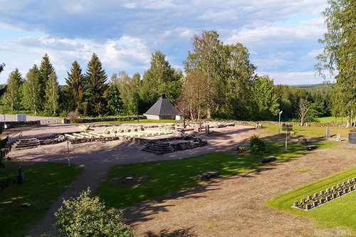 Kesäinen nurmialue, jota halkoo hiekkaisen kulkuväylät. Keskellä kivisiä, vanhan kirkon palamisen jäljiltä jääneitä kirkonpohjia, joiden keskellä pitkiä puupenkkejä ja alttari. Taustalla huoltorakennus sekä metsää.