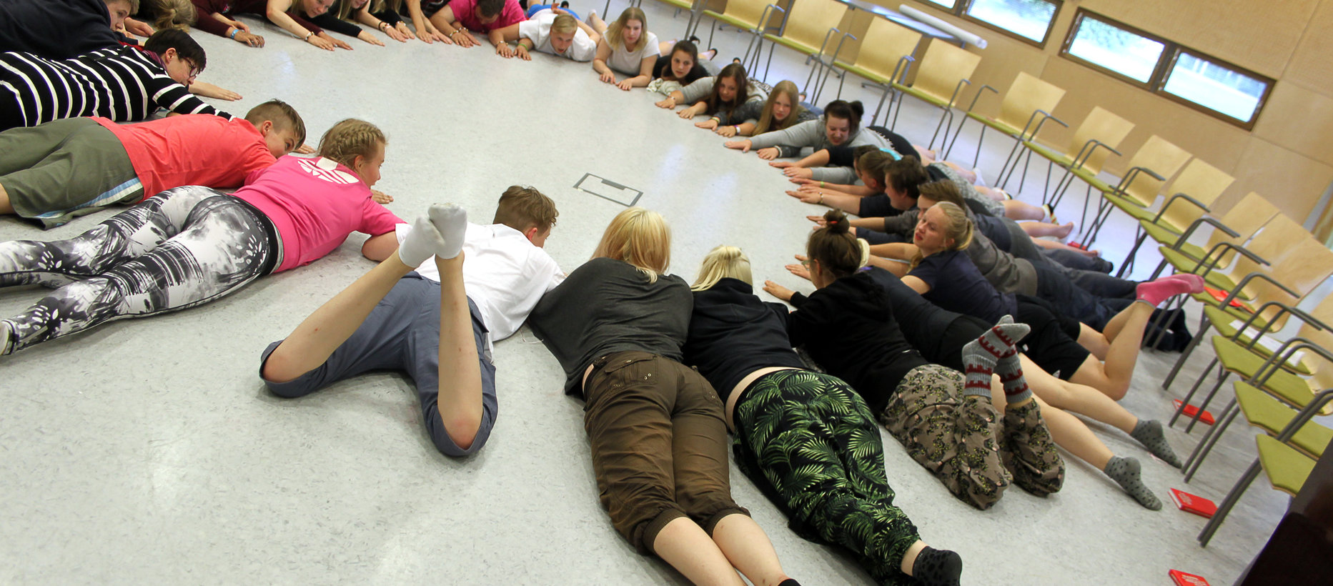 Joukko rippikoulunuoria makaa opetustilanteessa vatsallaan ringissä lattialla, kädet eteen ojennettuina.