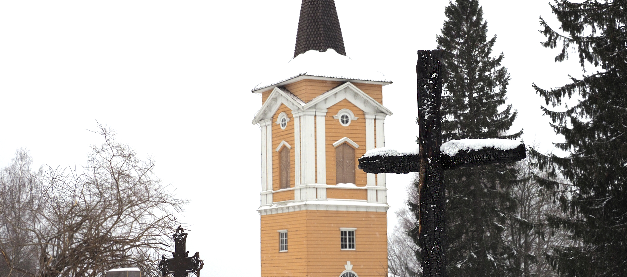Keltainen kellotapuli taustalla, lumisadetta ja palaneista puista tehty risti etualalla.
