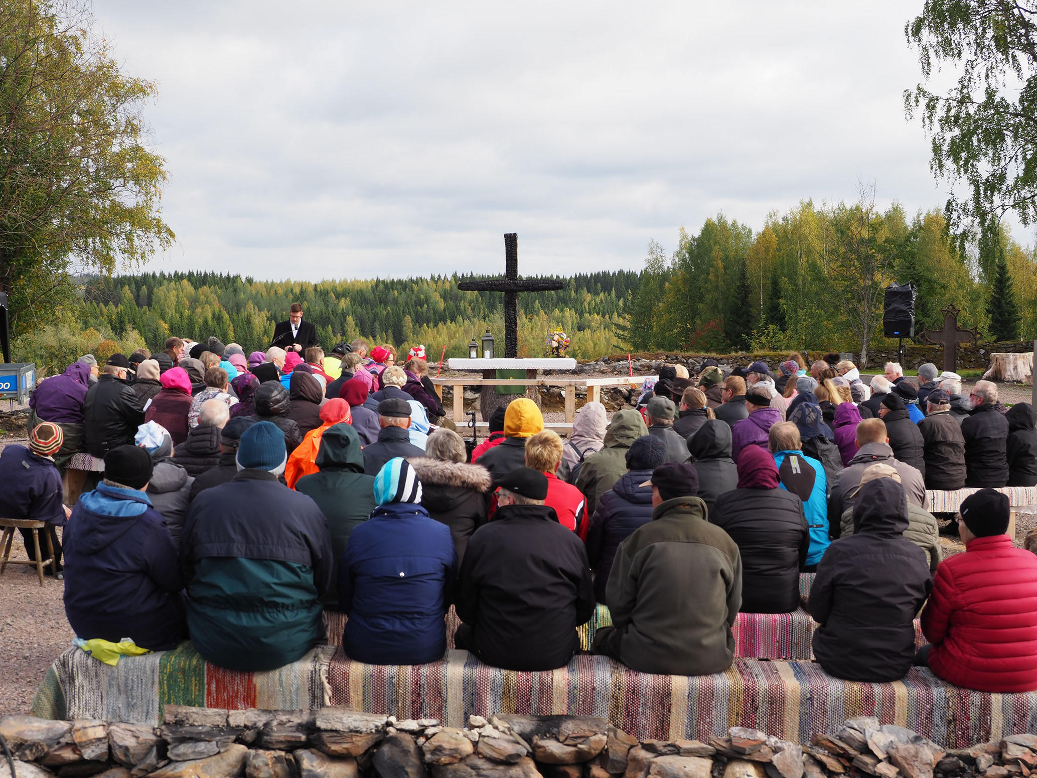 ihmisiä istumassa vieri vieressä penkeillä ulkona, etualalla risti