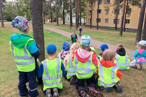 Kouluikäisiä huomioliivein puettuja lapsia kuvattuna selin puistossa jonglööraavaa ohjaajaa katsoen.