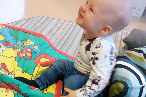 Vauva istuu lattialla värikkään leikkimaton päällä, katsoo ylös ja hymyilee.