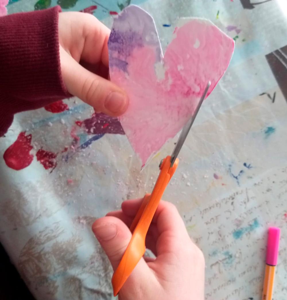 Lapsen käsi leikkaamassa saksilla vesivärein väritettyä paperia sydämen muotoiseksi.