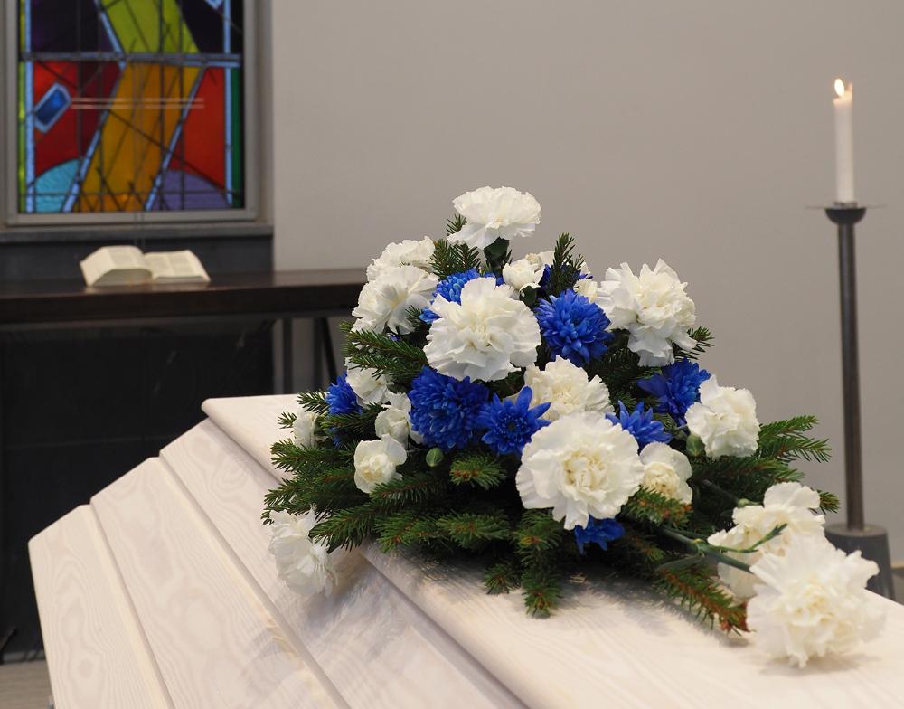 Valkoinen arkku, jonka päällä sinivalkoinen kukkakimppu, taustalla lasimaalausikkuna ja palava kynttilä.