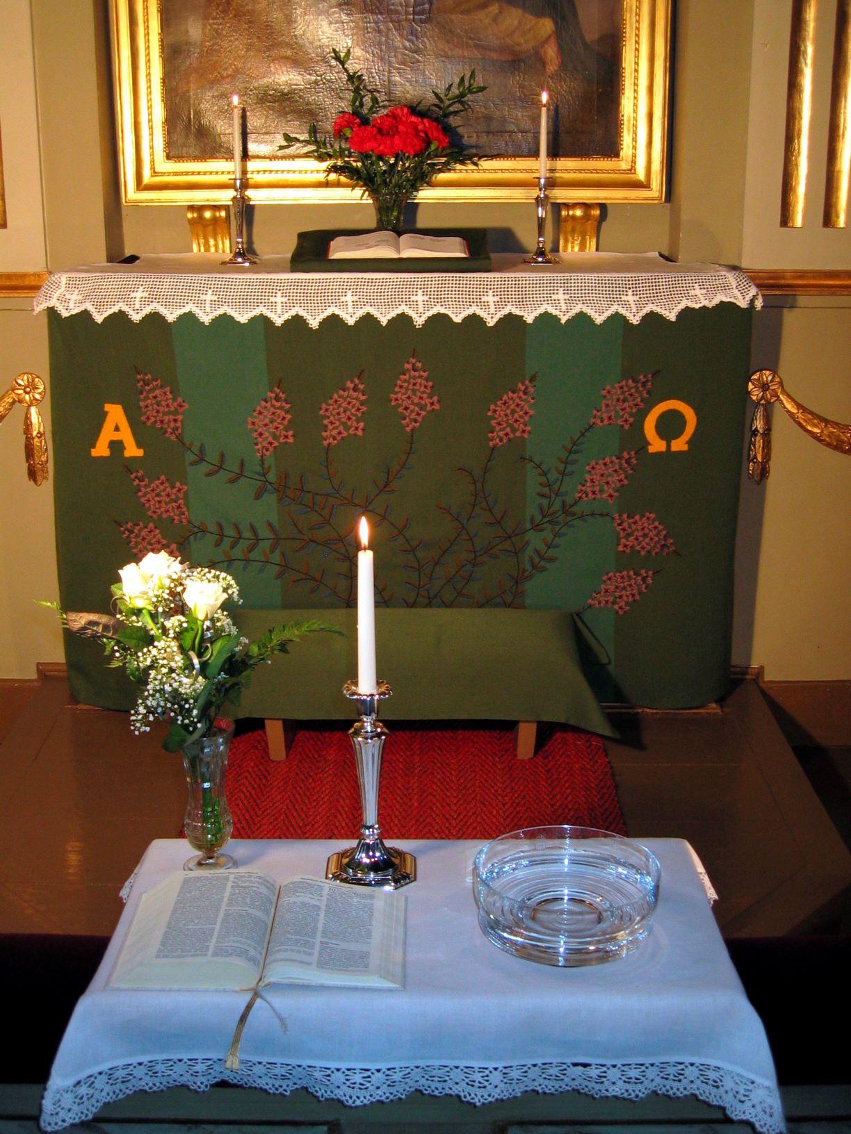 Kastepöytä, jossa kynttilä, kukka ja kastemalja.