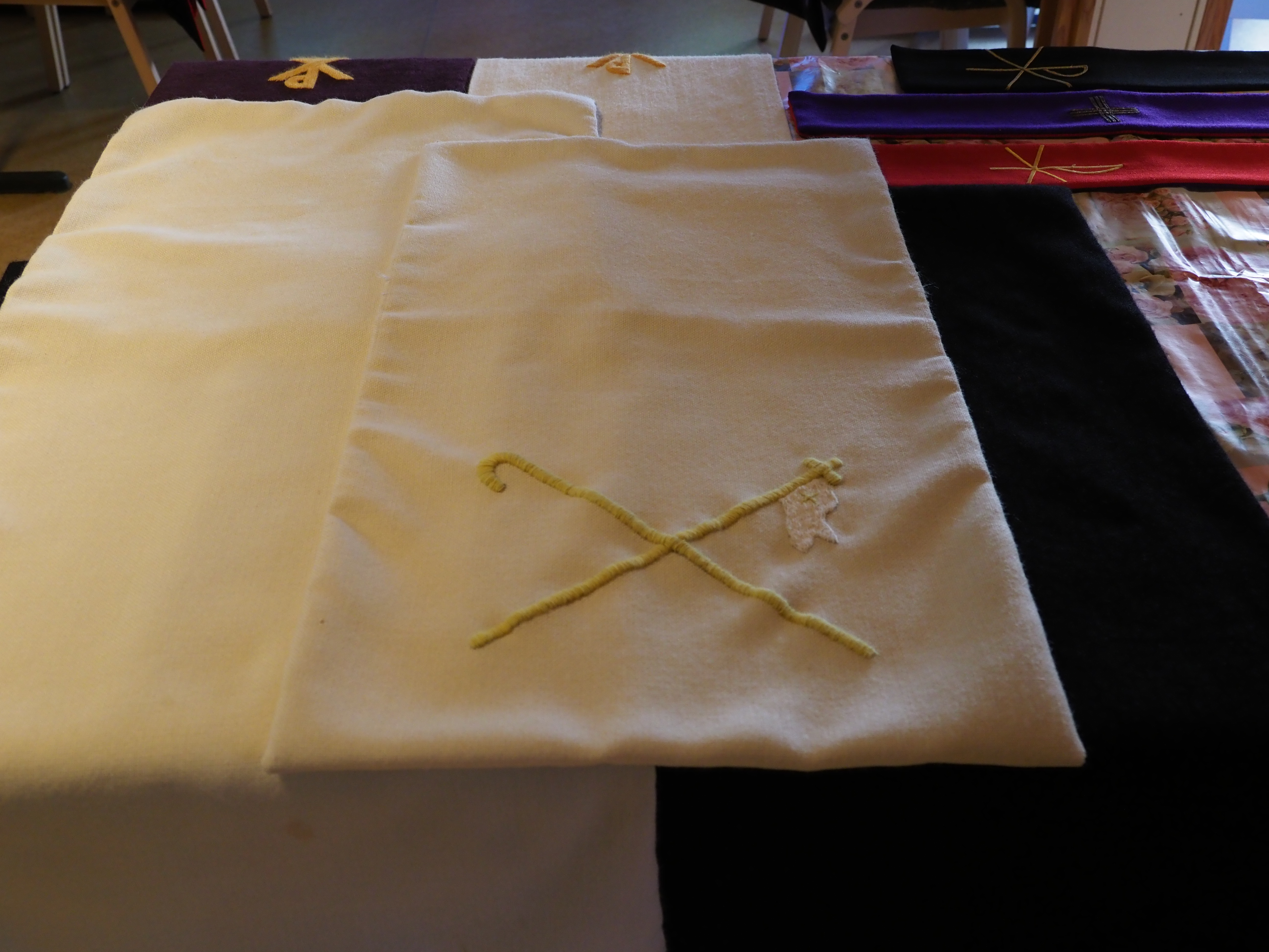 Edessä valkoisia kirkkotekstiilejä, joissa keltaista kirjailua. Takana eri värisiä tekstiilejä.