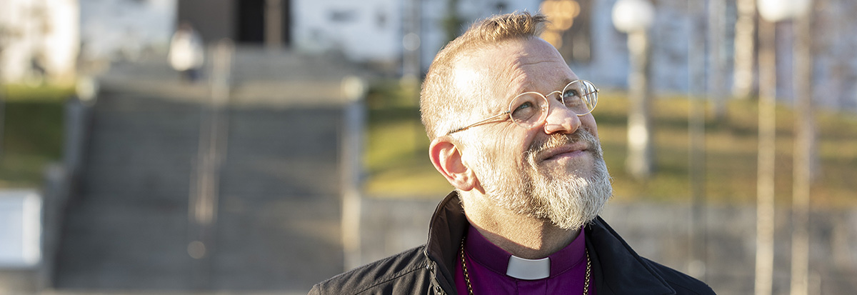 Mies ulkona kirkon edessä, violetissa piispan paidassa, katsoo ylös.