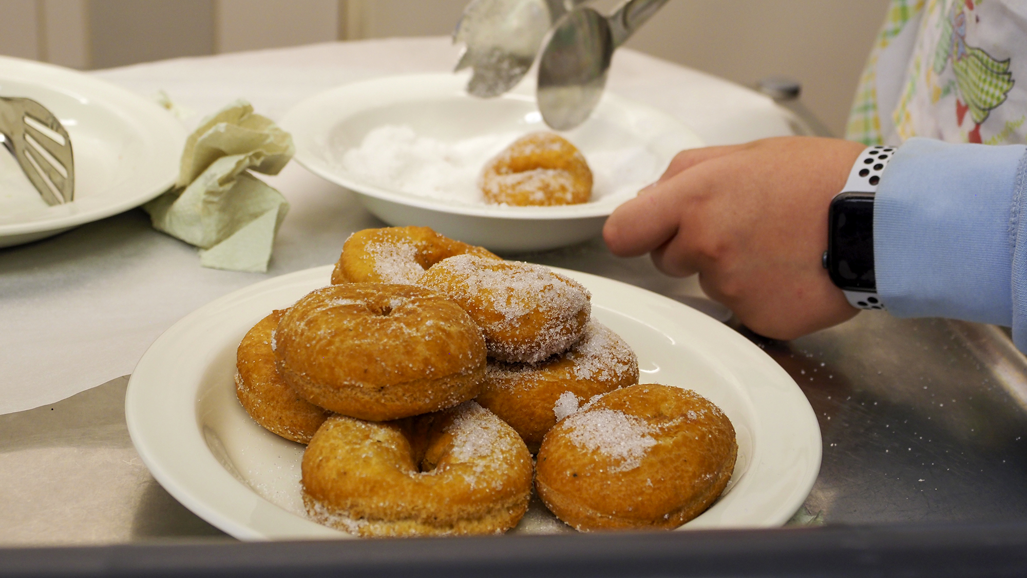 Lautasella on donitseja sokerikuorrutteella. Taustalla näkyvät pihdit ja donitsi lautasella, jolla on sokeria.