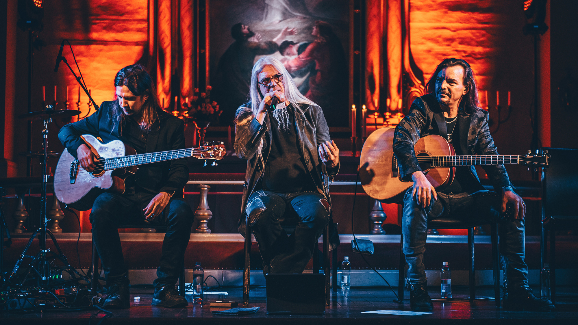 Marko Hietala istuu penkillä ja puhuu mikrofoniin. Vieressä istuu kaksi kitaristia.
