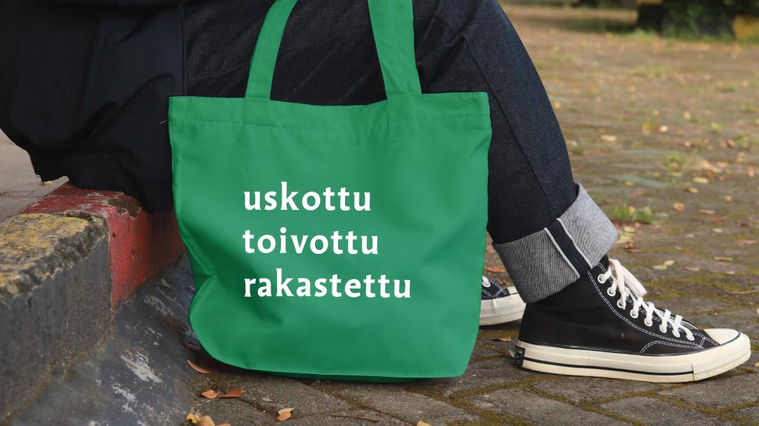 Vihreä kangaskassi, jossa teksti uskottu, toivottu, rakastettu katukynnyksellä istuvan henkilön jalkojen juuressa.